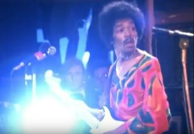 Jimi Hendrix on stage at Isle of Wight. To the right of Jimi's Stratocaster headstock Hendrix road crew member Howard Parker, aka "H," observes the troubled set.